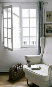 Sillón blanco frente a una ventana abierta en una habitación luminosa y serena. En el apoyabrazos hay una taza y un libro. Todo el ambiente transmite calma, aire fresco y pausa cotidiana.