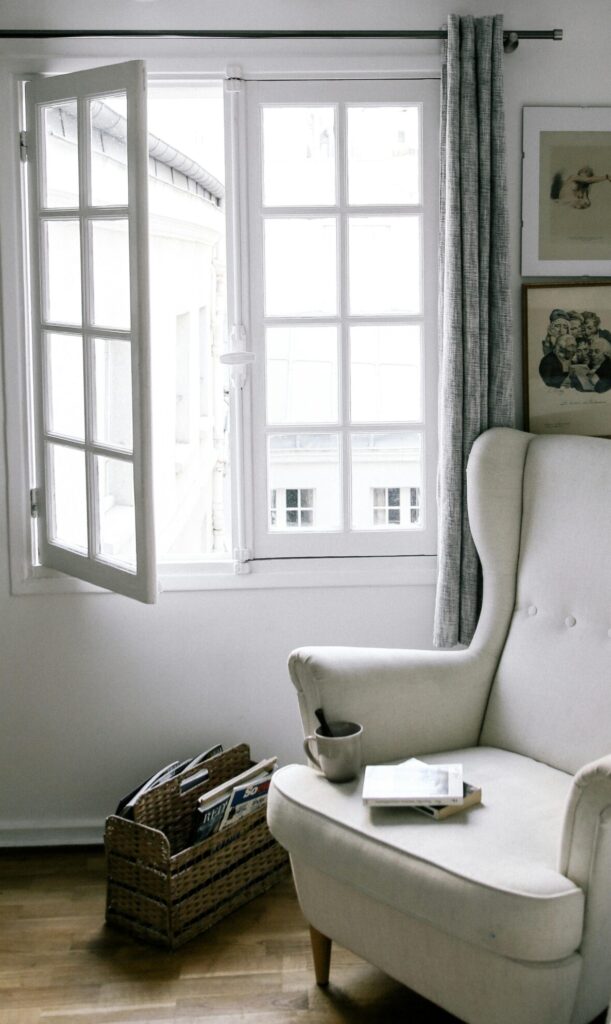 Sillón blanco frente a una ventana abierta en una habitación luminosa y serena. En el apoyabrazos hay una taza y un libro. Todo el ambiente transmite calma, aire fresco y pausa cotidiana.
